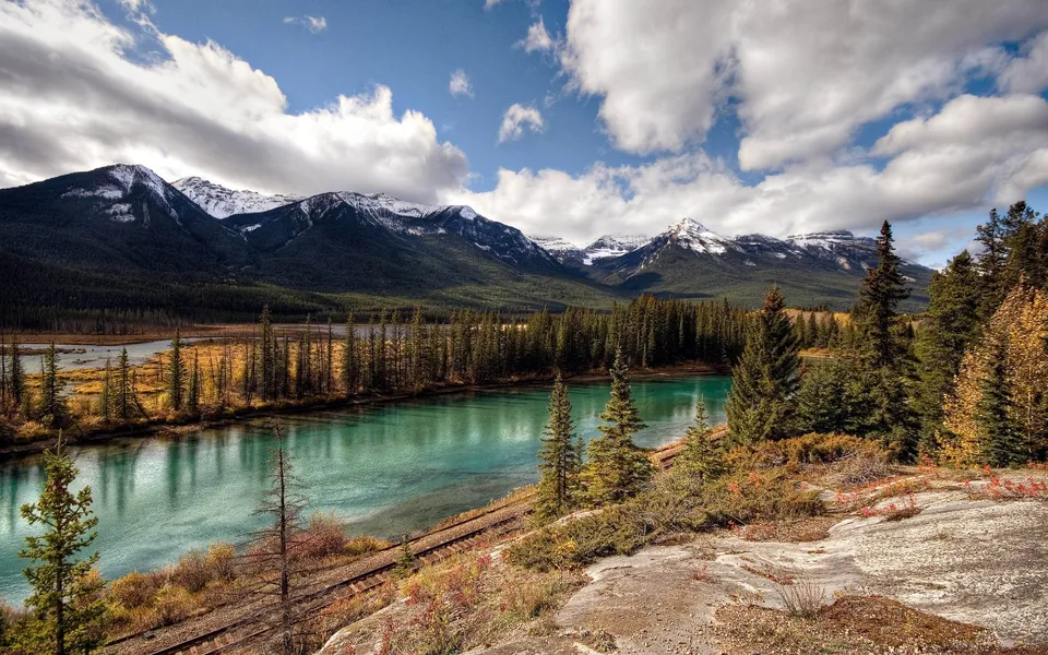A wide, turquoise river flows past snow-capped mountains and evergreen forests under a cloudy sky.