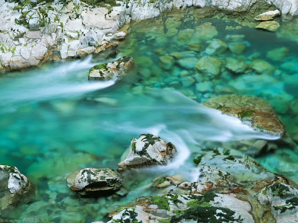 A clear, turquoise mountain stream flows over rocks covered in moss and lichen.