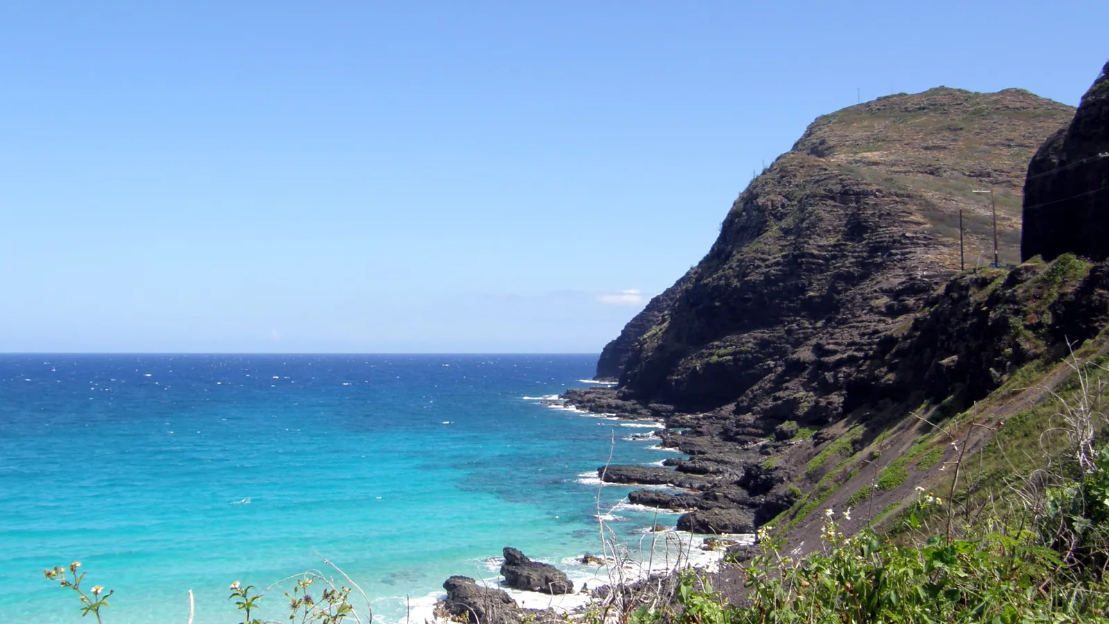 Turquoise ocean waters meet a rugged, dark volcanic coastline under a clear blue sky.