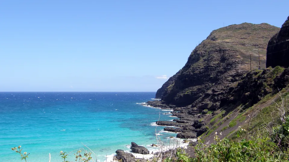 Turquoise ocean waters meet a rugged, dark volcanic coastline under a clear blue sky.