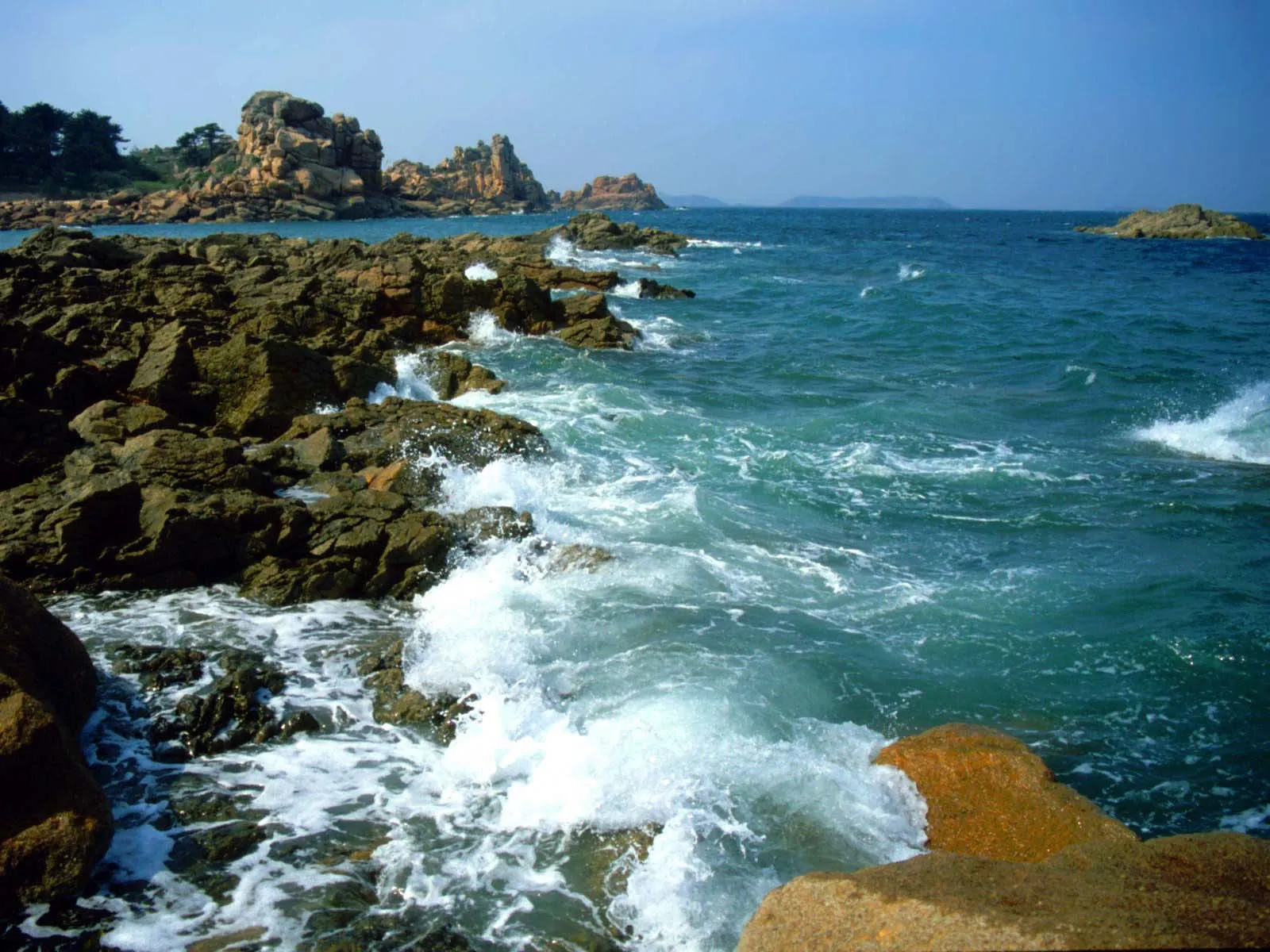 Turquoise ocean waves break against a dark, rocky shore in bright daylight.