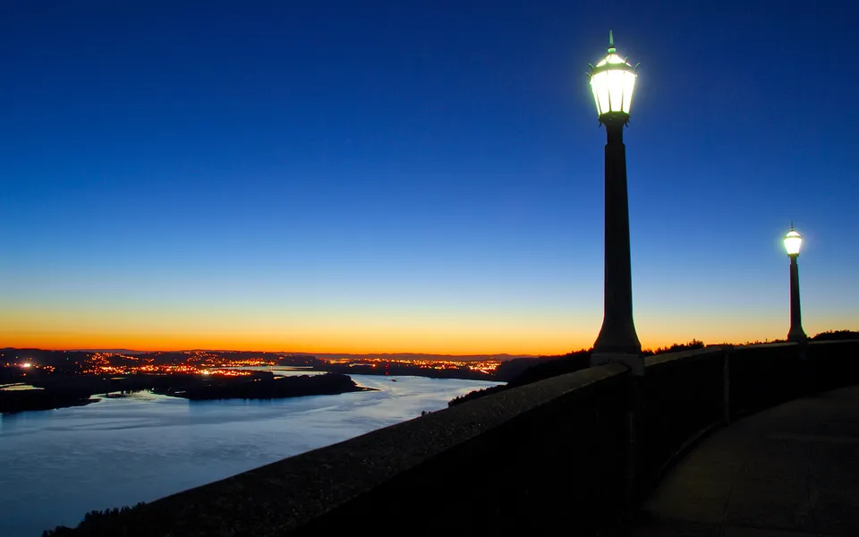 A wide river flows past a city illuminated by lights at twilight, with two vintage lampposts in the foreground.