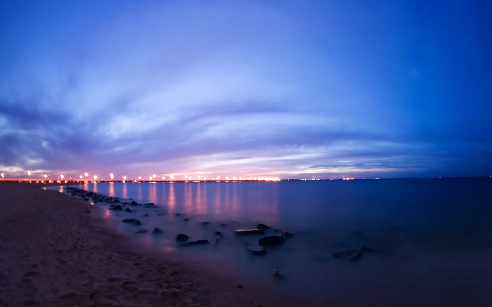 A long exposure shot of a beach at twilight with lights reflecting on the calm sea.