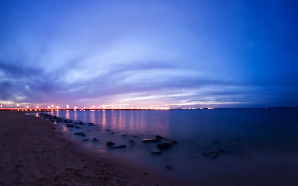A long exposure shot of a beach at twilight with lights reflecting on the calm sea.