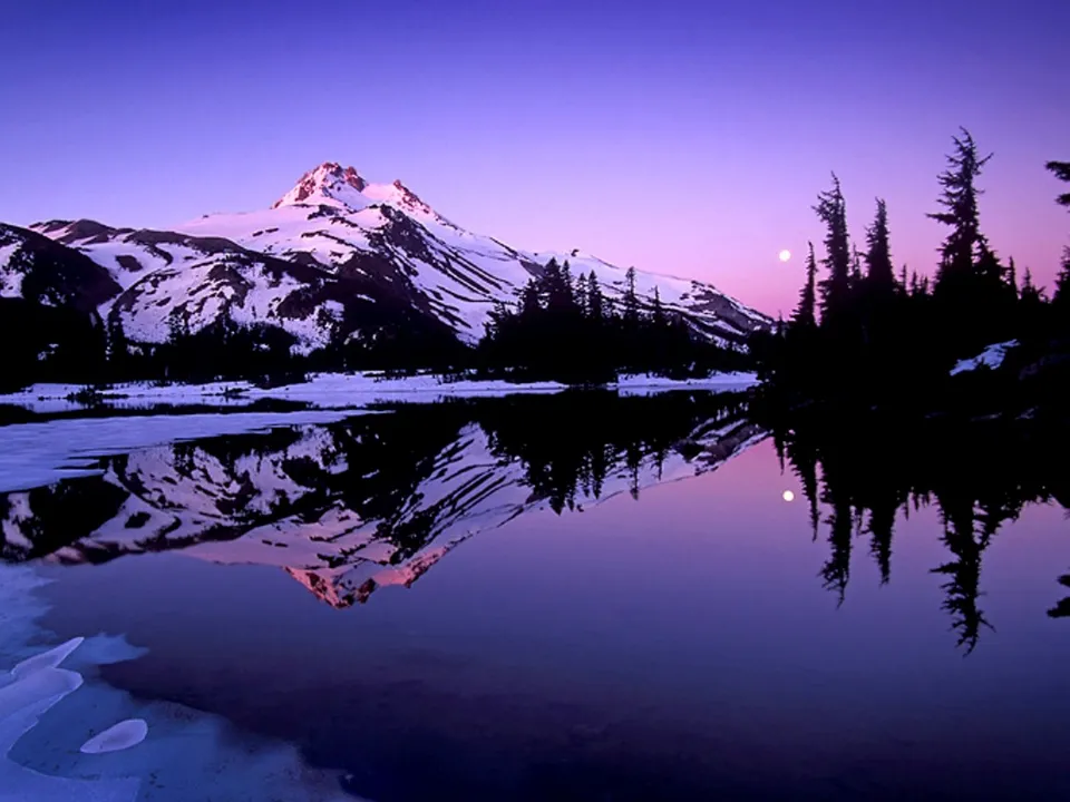 Snowy mountain peaks are reflected in a lake at twilight with purple and pink skies.