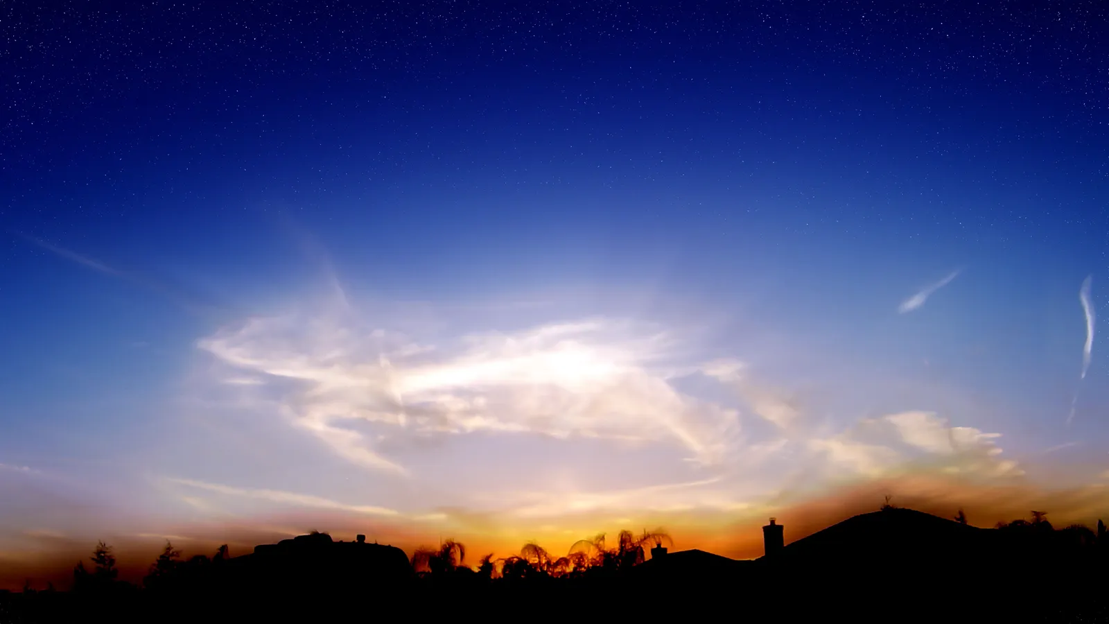 A vibrant twilight sky with stars visible above a dark silhouette of houses and trees.