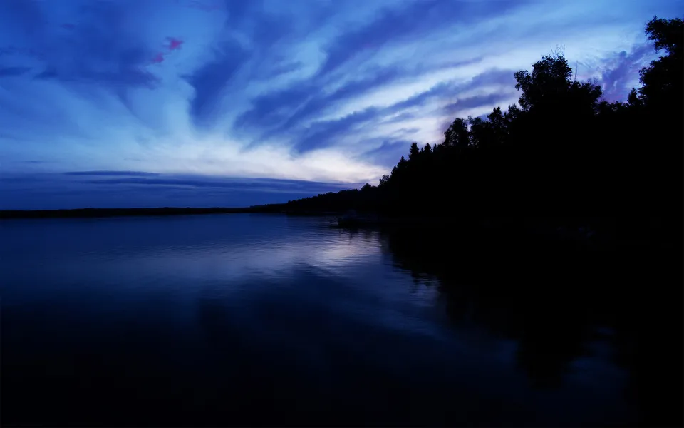 A dramatic blue twilight sky with wispy clouds reflects on the dark, calm surface of a lake.
