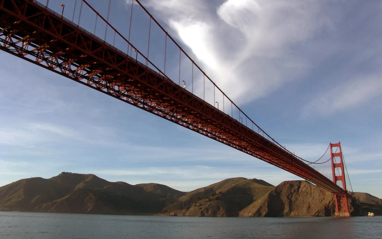 View from below the Golden Gate Bridge, showing its structure against a sky with wispy clouds and distant hills.