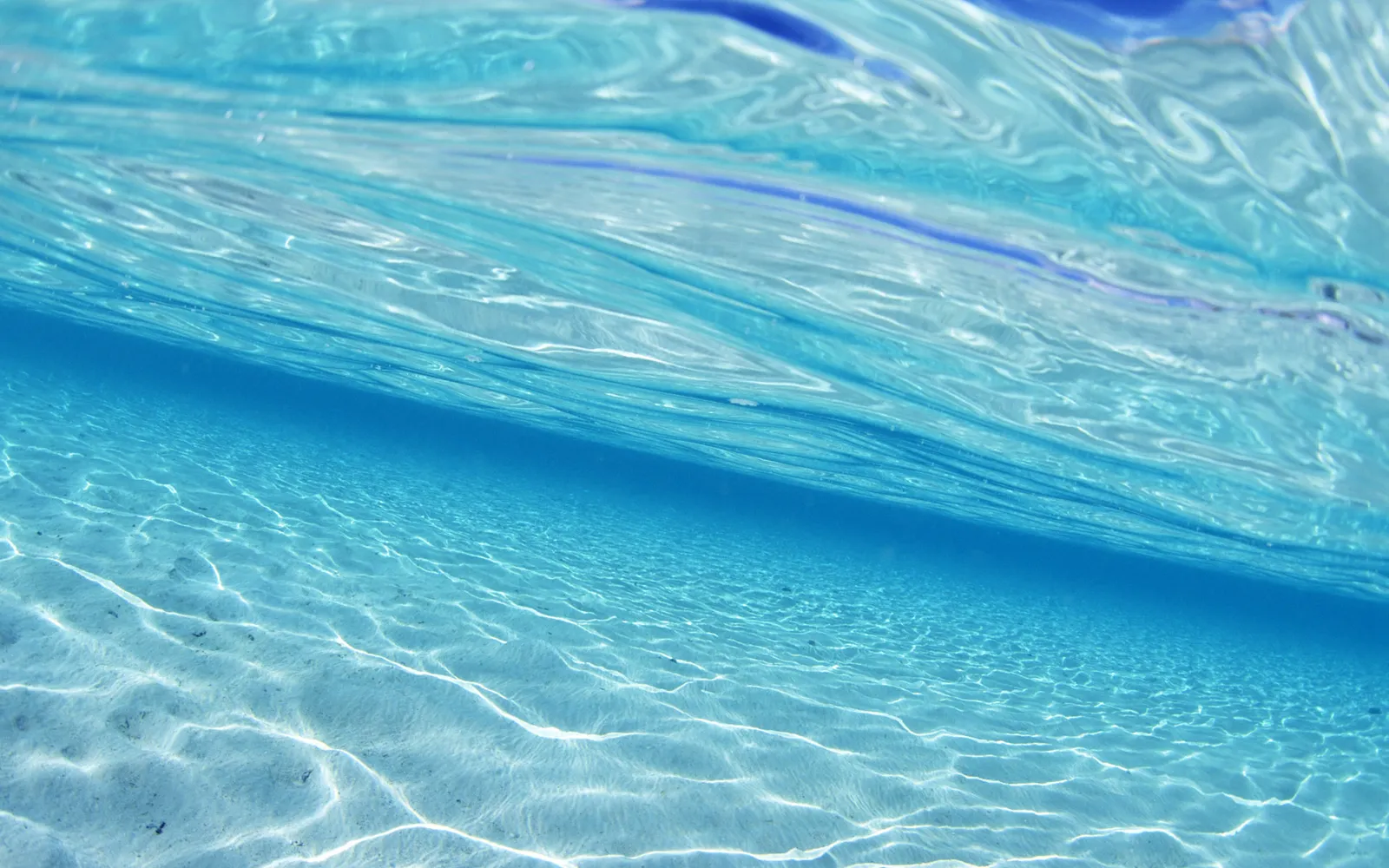 Underwater view of a sandy ocean floor with clear blue water and sunlight patterns on the sand.