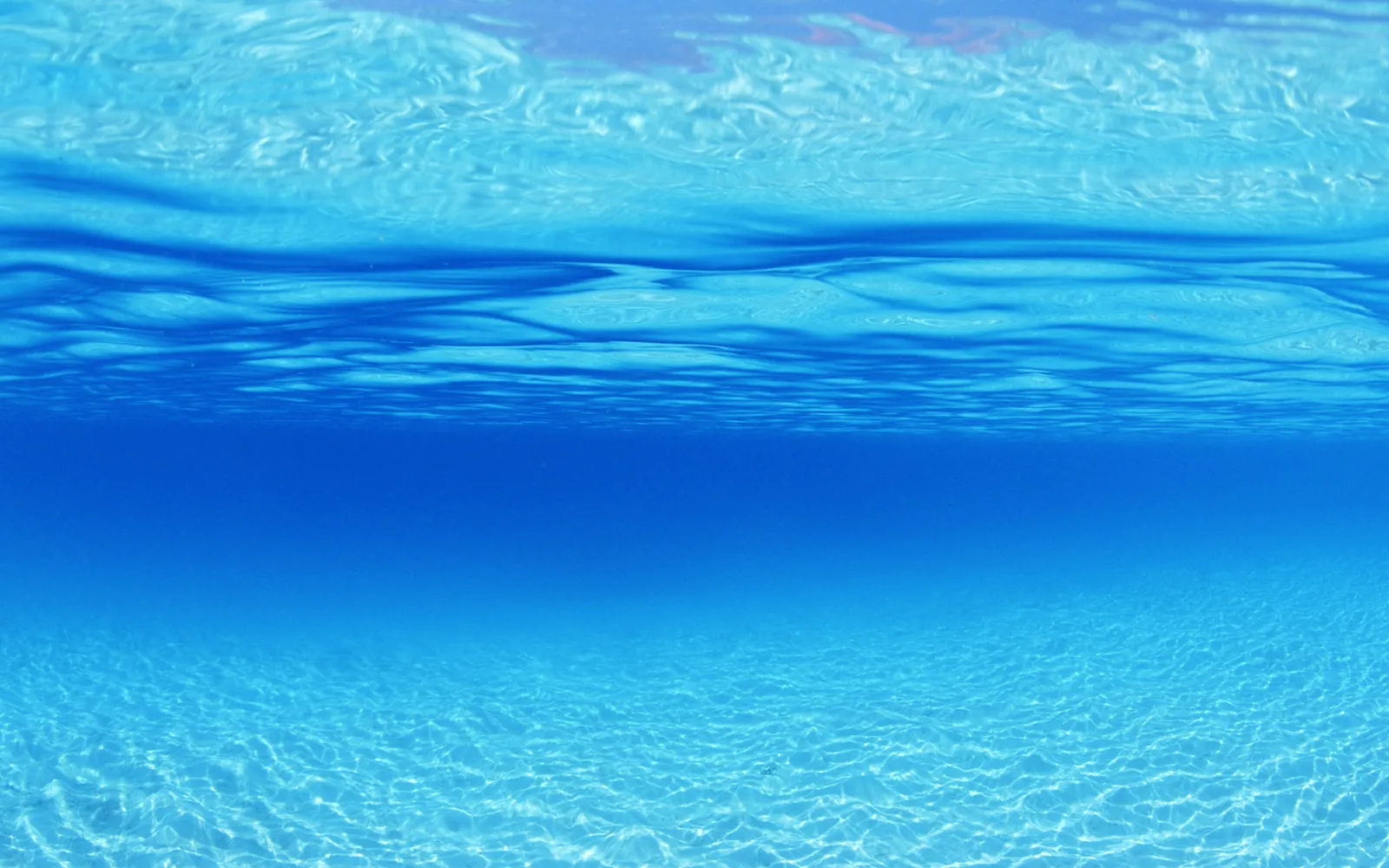 View from underwater looking up towards the surface, with sunlight filtering through the clear blue water onto a sandy seabed.