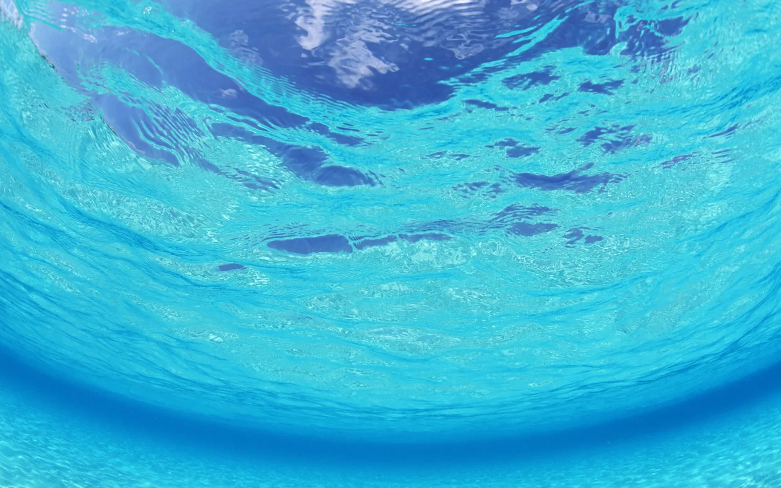 View from underwater looking up at the sunlit surface of clear blue ocean water with clouds visible.