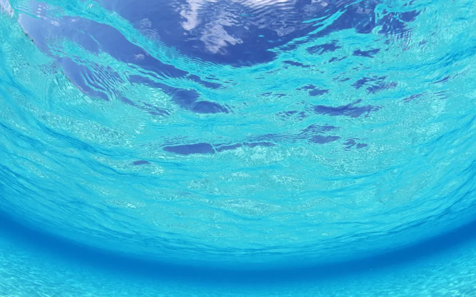 View from underwater looking up at the sunlit surface of clear blue ocean water with clouds visible.