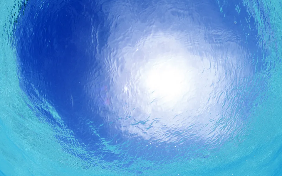 View from underwater looking up at the bright sun and blue sky through rippling water.