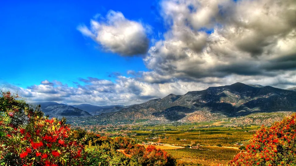 A wide valley with agricultural fields and distant mountains under a dramatic sky with clouds.