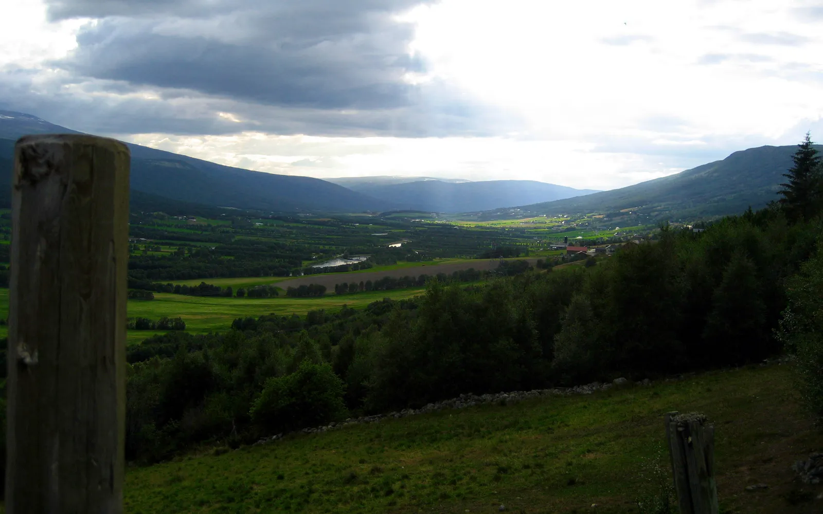 A wide valley with rolling green hills, trees, and a distant lake, under a dramatic cloudy sky with sunbeams.