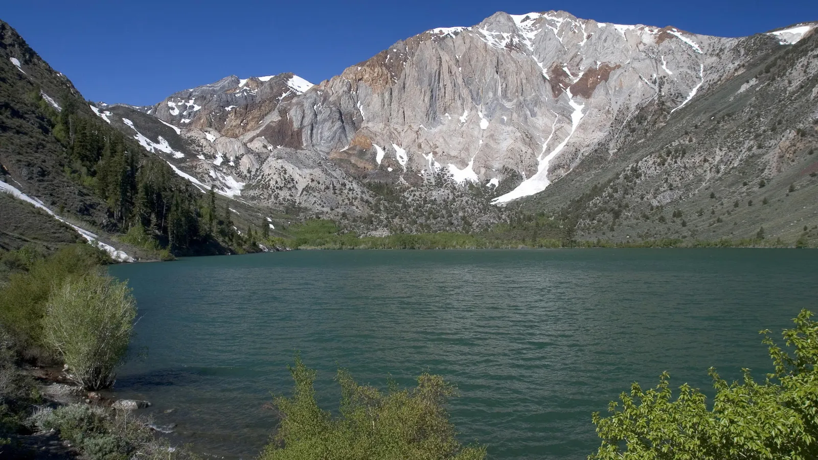 A wide, deep blue lake sits at the base of rugged, snow-capped mountains under a clear blue sky.
