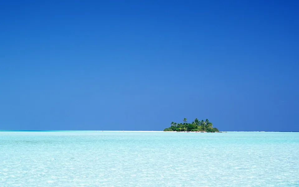 A small, uninhabited island with palm trees sits in the middle of a vast, clear turquoise ocean under a cloudless blue sky.