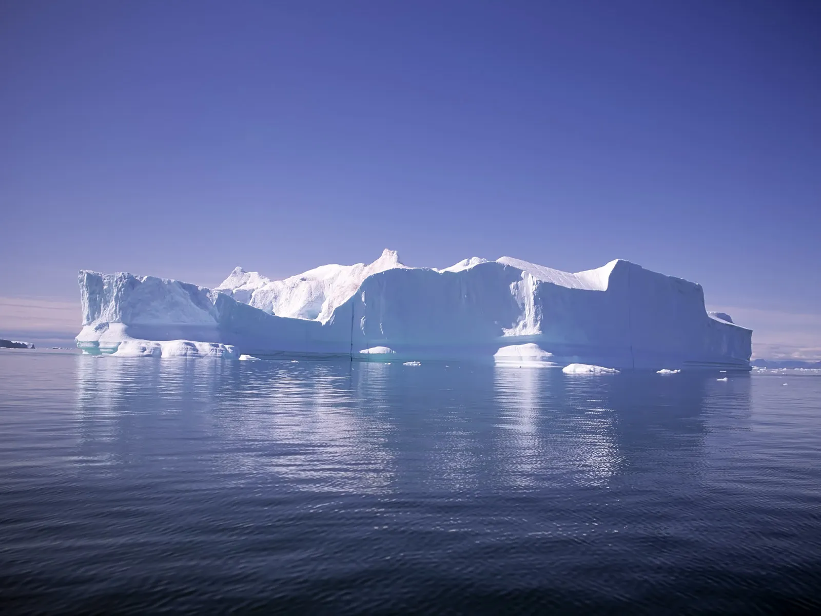 A large, sculpted iceberg with blue icy surfaces reflects in deep blue water under a bright sky.