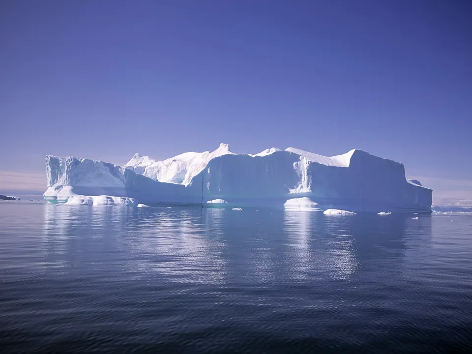 A large, sculpted iceberg with blue icy surfaces reflects in deep blue water under a bright sky.