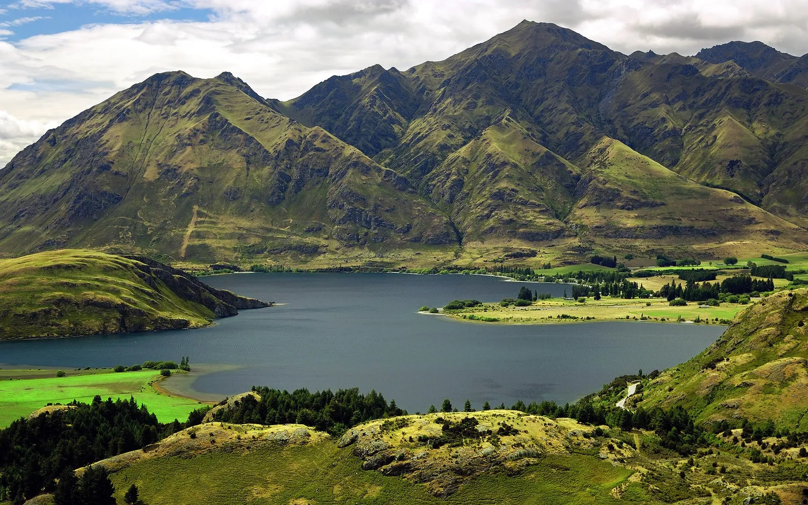 A dark lake is surrounded by rolling green hills and rugged mountains under a cloudy sky.