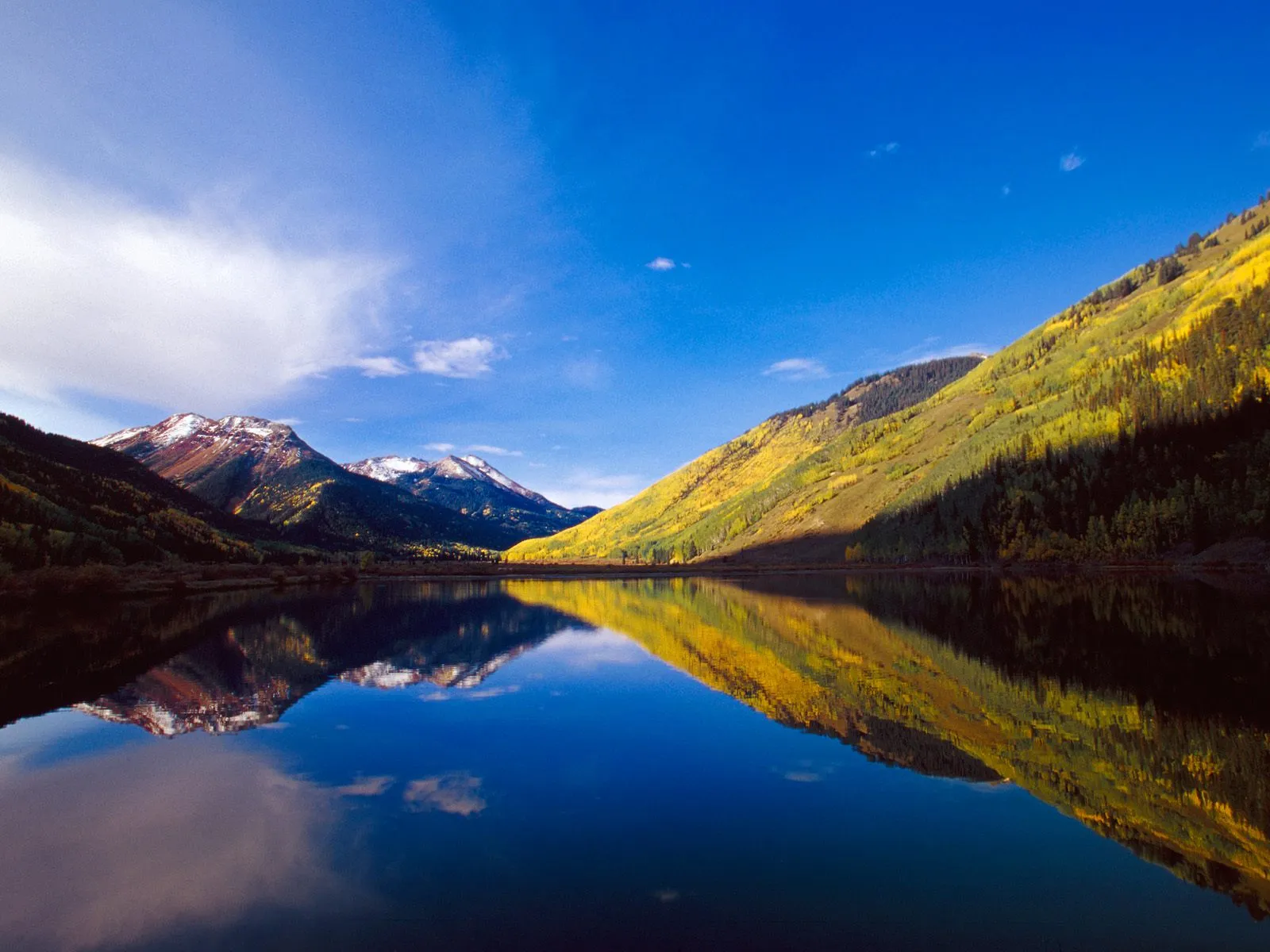 A mountain lake reflects vibrant autumn hills and snow-dusted peaks under a blue sky.