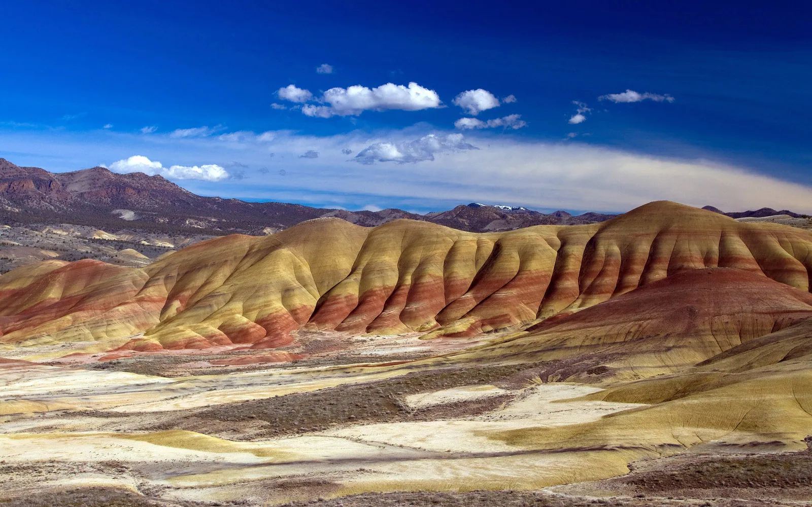 The Painted Hills in Oregon display distinct layers of colorful soil under a bright blue sky with scattered clouds.