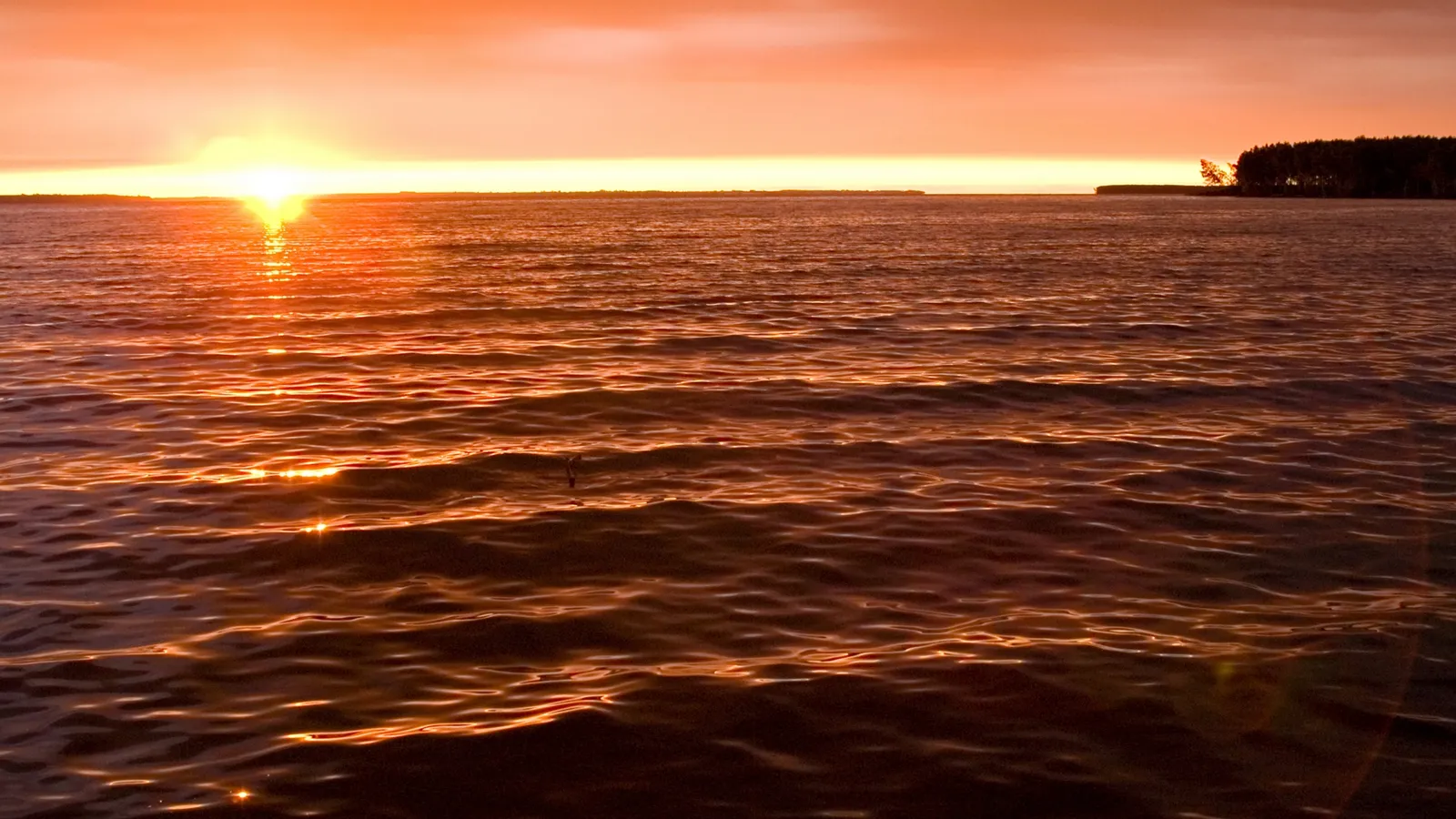 The sun sets over a wide, calm ocean, casting vibrant orange hues across the sky and water.