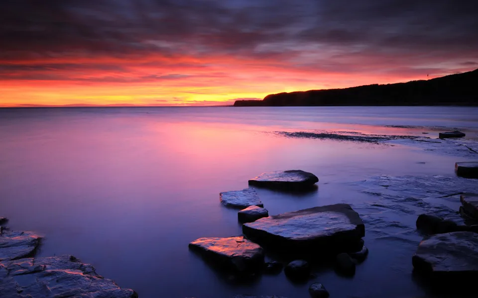 A vibrant sunset with pink and orange hues over a calm ocean with rocks in the foreground.