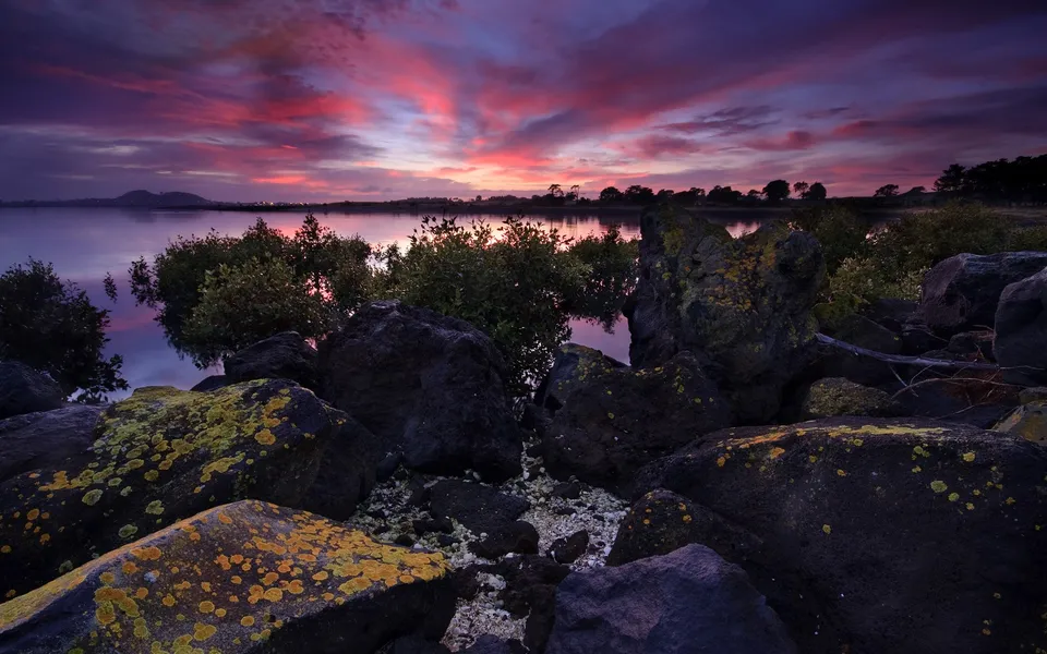 A vibrant sunset with pink and purple clouds reflects in calm water, with lichen-covered rocks in the foreground.