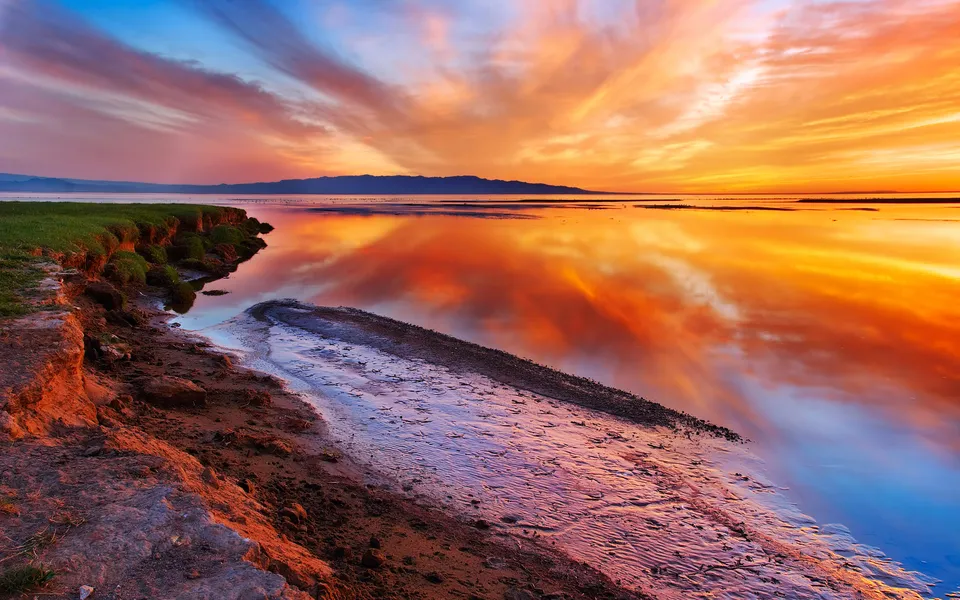 A dramatic sunset with fiery orange and pink clouds reflecting on a calm body of water, with a grassy cliff in the foreground.