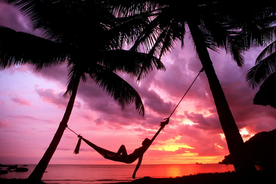 Silhouette of a person in a hammock between palm trees at sunset with a vibrant sky.