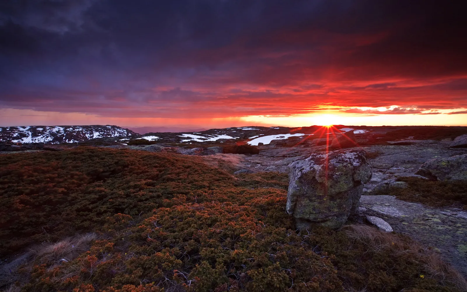 A dramatic sunset with intense orange and red hues over a rocky landscape with patches of snow.