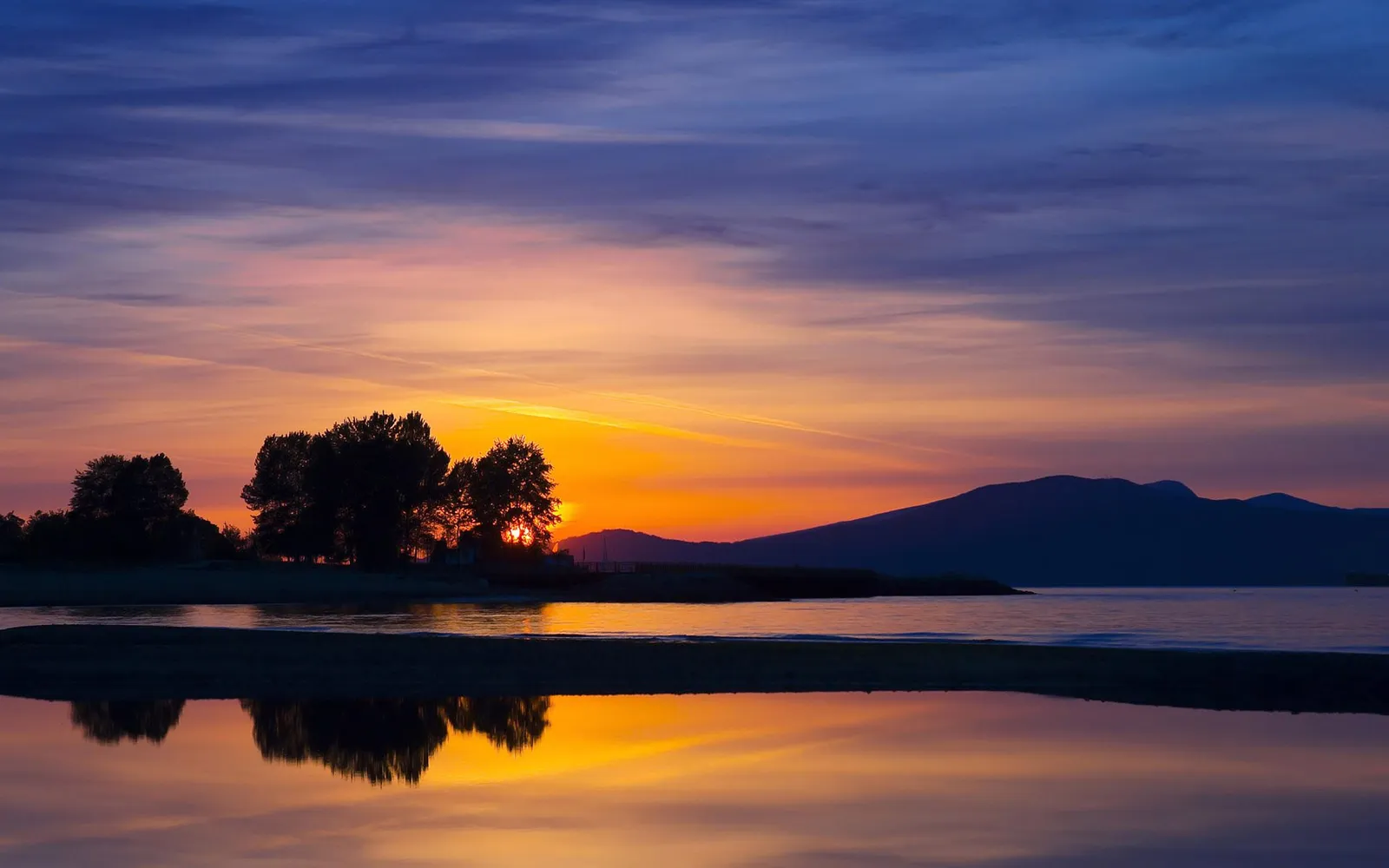 A vibrant sunset paints the sky with orange, purple, and blue hues over a calm body of water with trees.