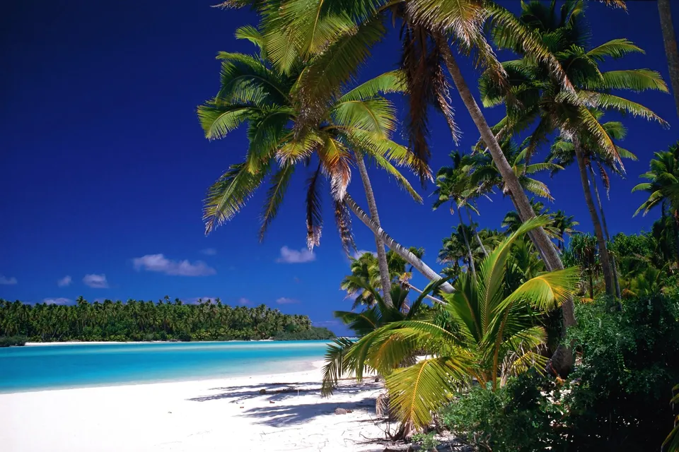 A bright tropical beach with white sand, palm trees, and clear turquoise water under a deep blue sky.