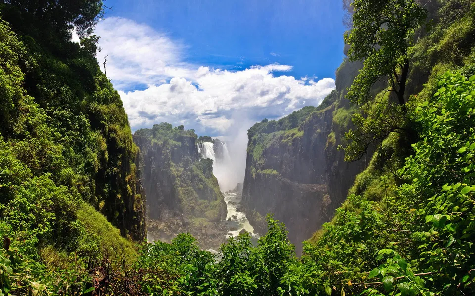 A wide view of Victoria Falls with mist rising, framed by steep, green cliffs and lush vegetation.