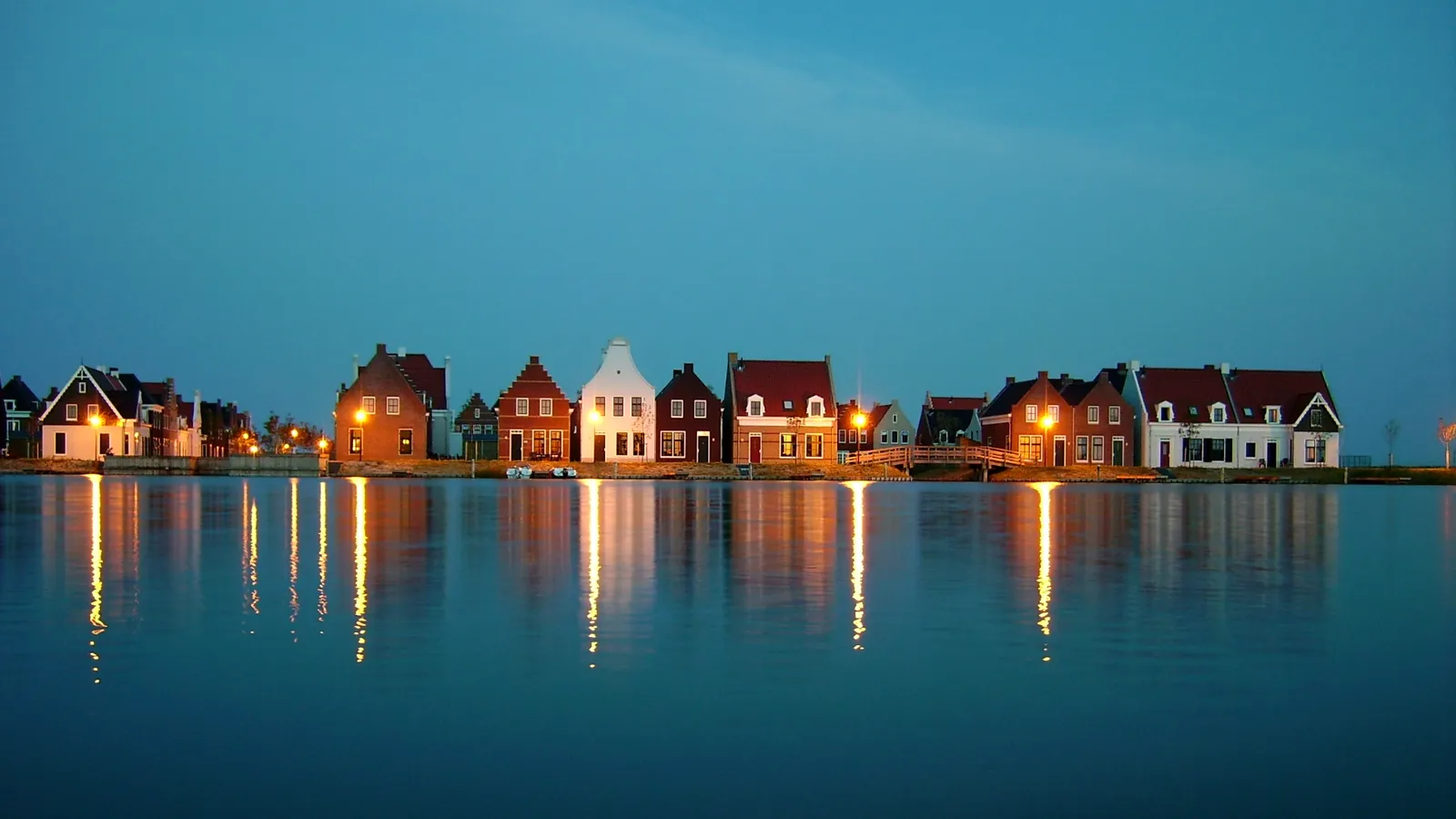 A row of traditional Dutch houses with lights on, reflected in calm water under a twilight sky.