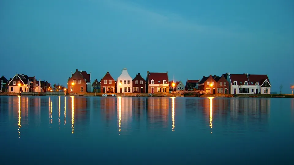 A row of traditional Dutch houses with lights on, reflected in calm water under a twilight sky.