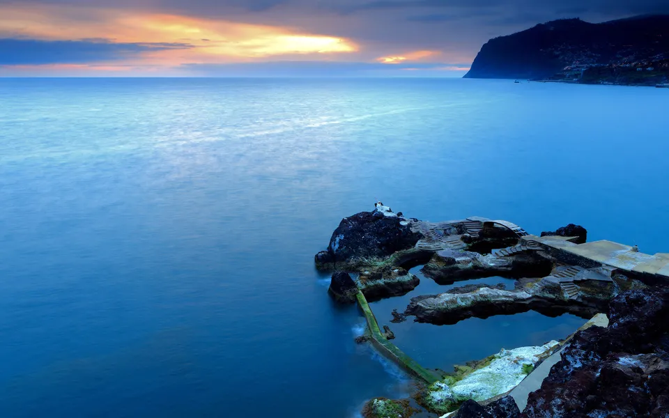 Natural volcanic rock pools on a coastline with the ocean and a colorful sunset sky in the background.