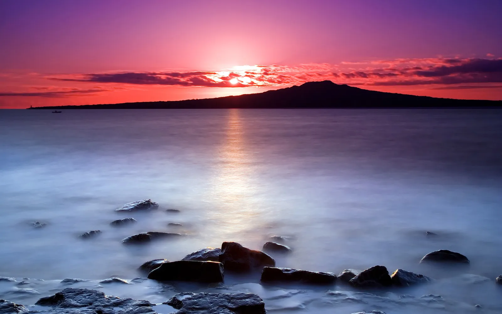 A silhouetted volcanic island is visible on the horizon during a vibrant sunset over a calm ocean.