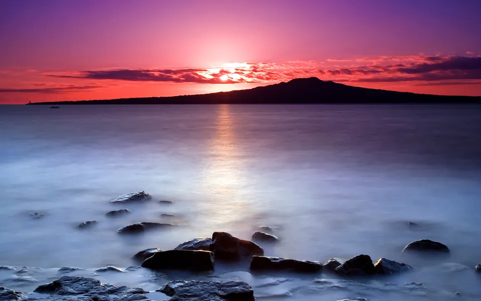 A silhouetted volcanic island is visible on the horizon during a vibrant sunset over a calm ocean.