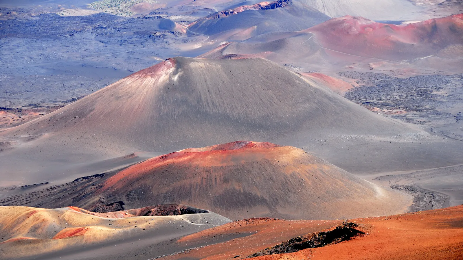 A vast, barren landscape of volcanic craters with slopes of red, orange, and gray hues under a bright sky.