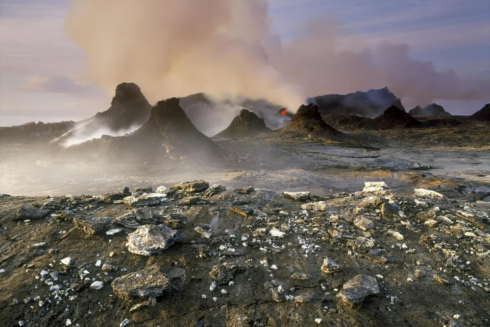 A volcanic landscape with active eruptions, smoke plumes, and glowing lava visible.