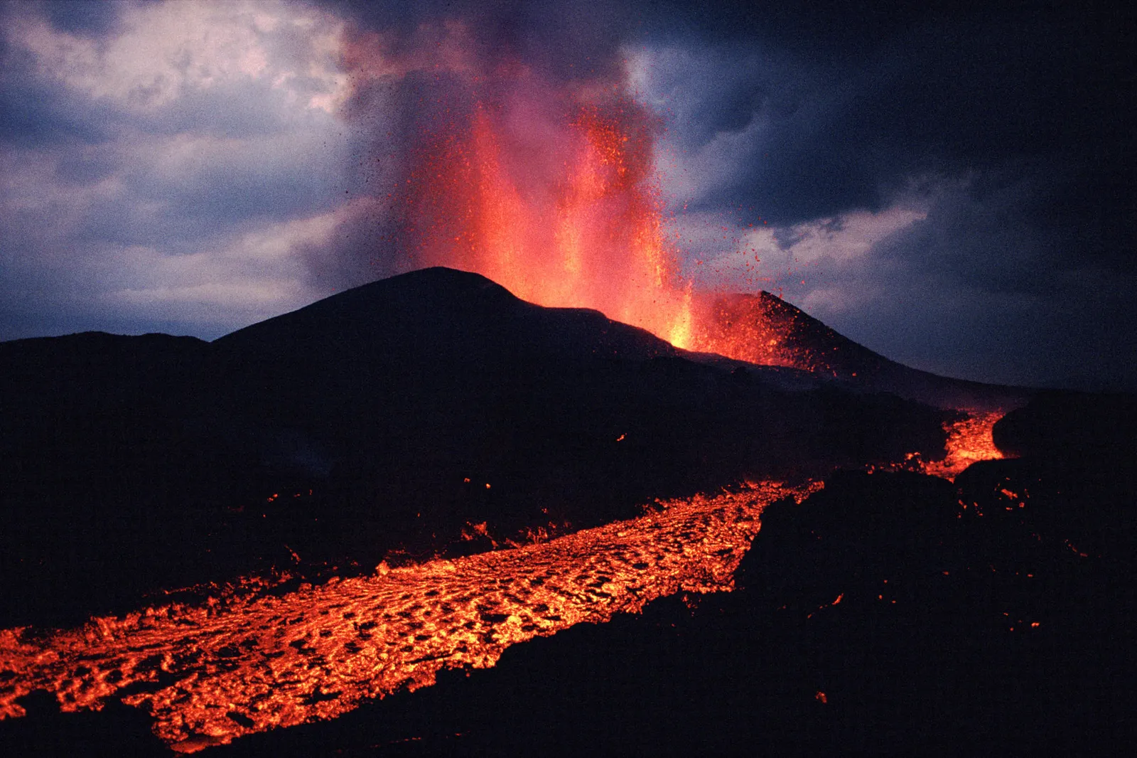 A volcano erupts with bright orange lava spewing into the dark, cloudy sky, with a river of lava flowing down the mountainside.