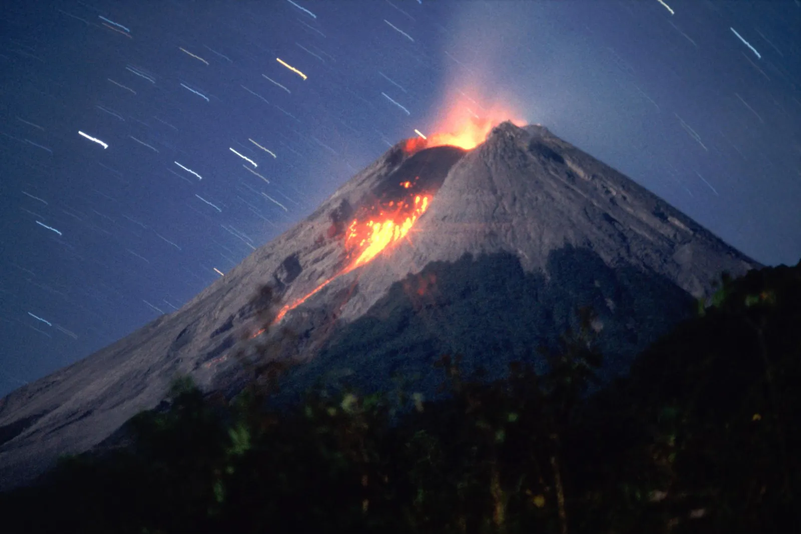 A volcano erupts with lava flowing down its side under a dark blue sky with star trails.