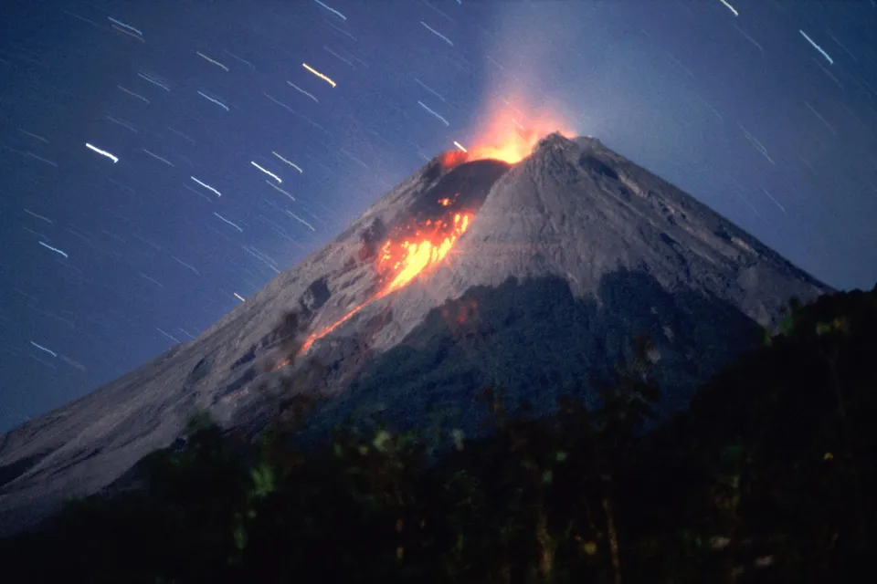 A volcano erupts with lava flowing down its side under a dark blue sky with star trails.