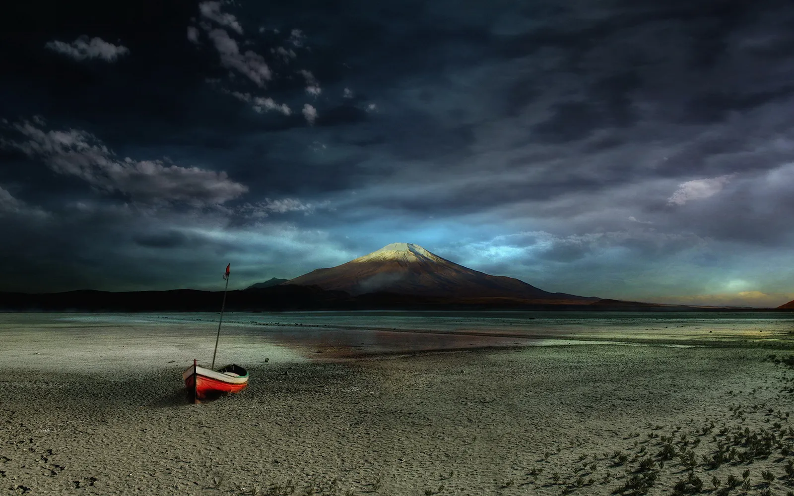A red and white boat sits on a dry, cracked lake bed with a large volcano in the background under a dramatic sky.