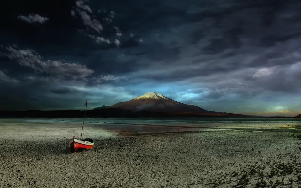 A red and white boat sits on a dry, cracked lake bed with a large volcano in the background under a dramatic sky.