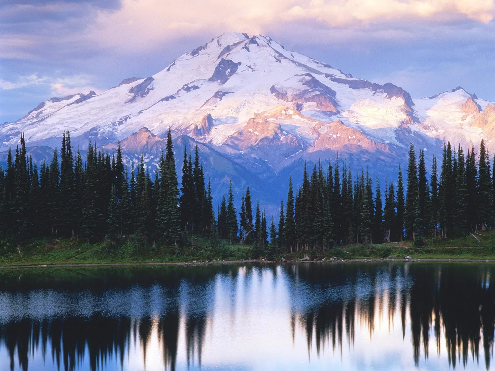 Snow-covered mountain peaks illuminated by warm light, reflected in a calm lake with evergreen trees in the foreground.