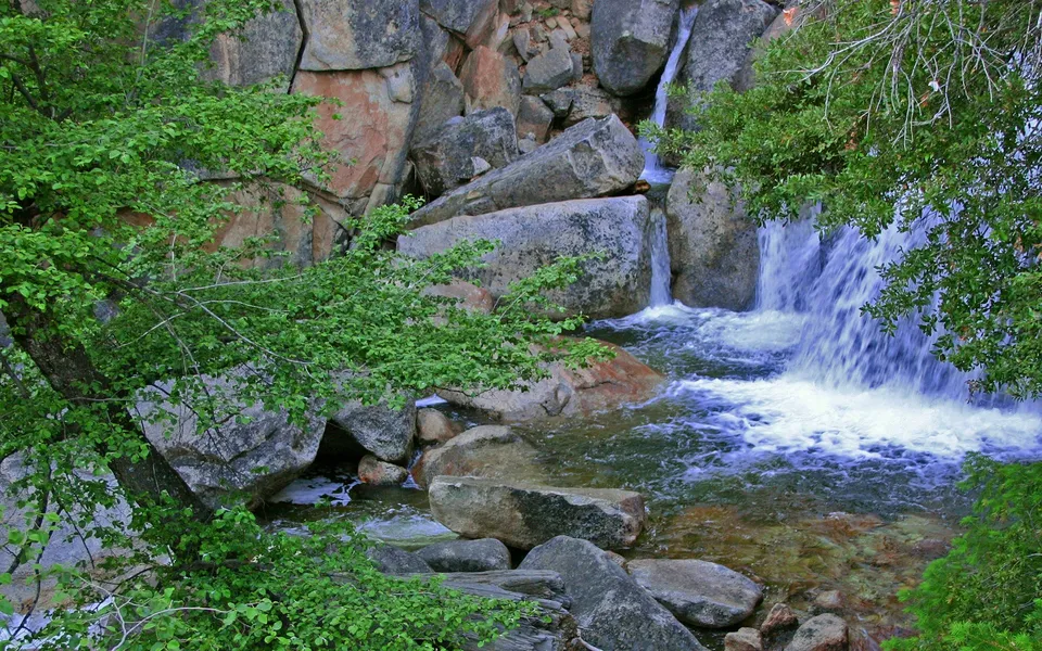 A cascading waterfall flows over large, smooth boulders surrounded by lush green trees.