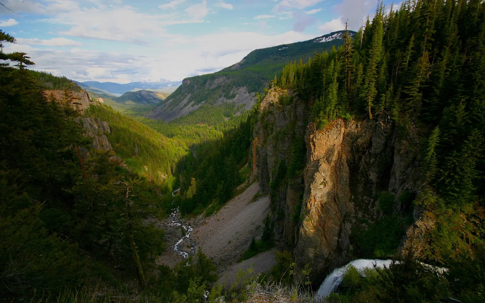 A powerful waterfall cascades down a rocky cliff face into a verdant, tree-filled canyon.